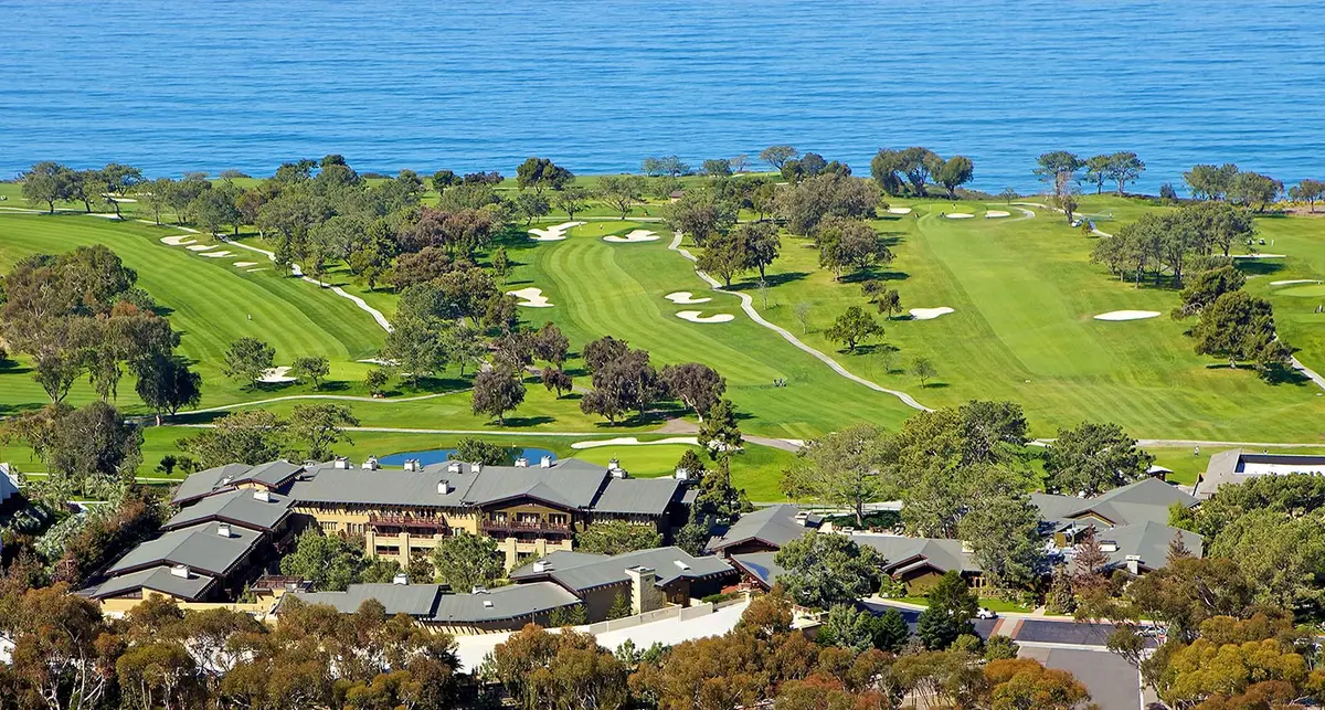 An aerial view of a low timber lodge beside a green golf course on a cliff above the Pacific