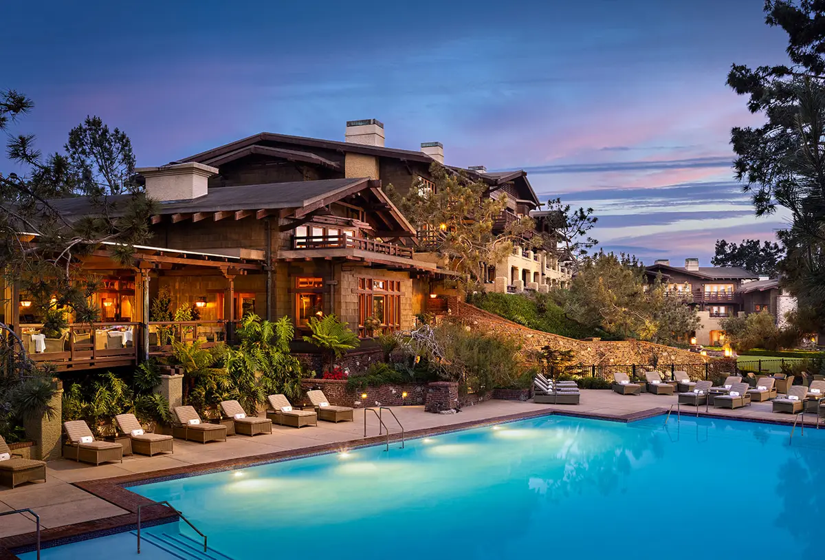 A timber and stone craftsman lodge lit from inside at dusk above a lit swimming pool