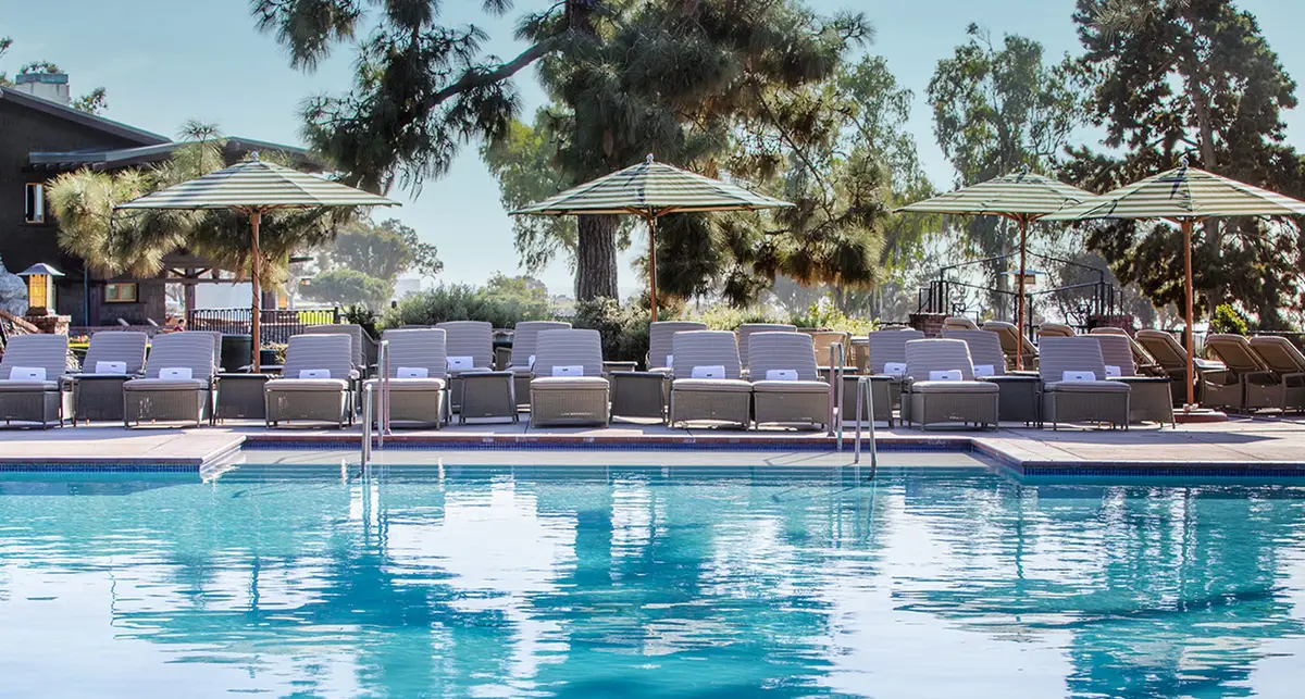 A pool with rows of loungers and cream umbrellas under tall pines