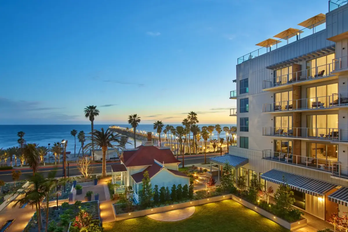 A glass and timber resort lit at dusk with lawns, palms and the beach beyond
