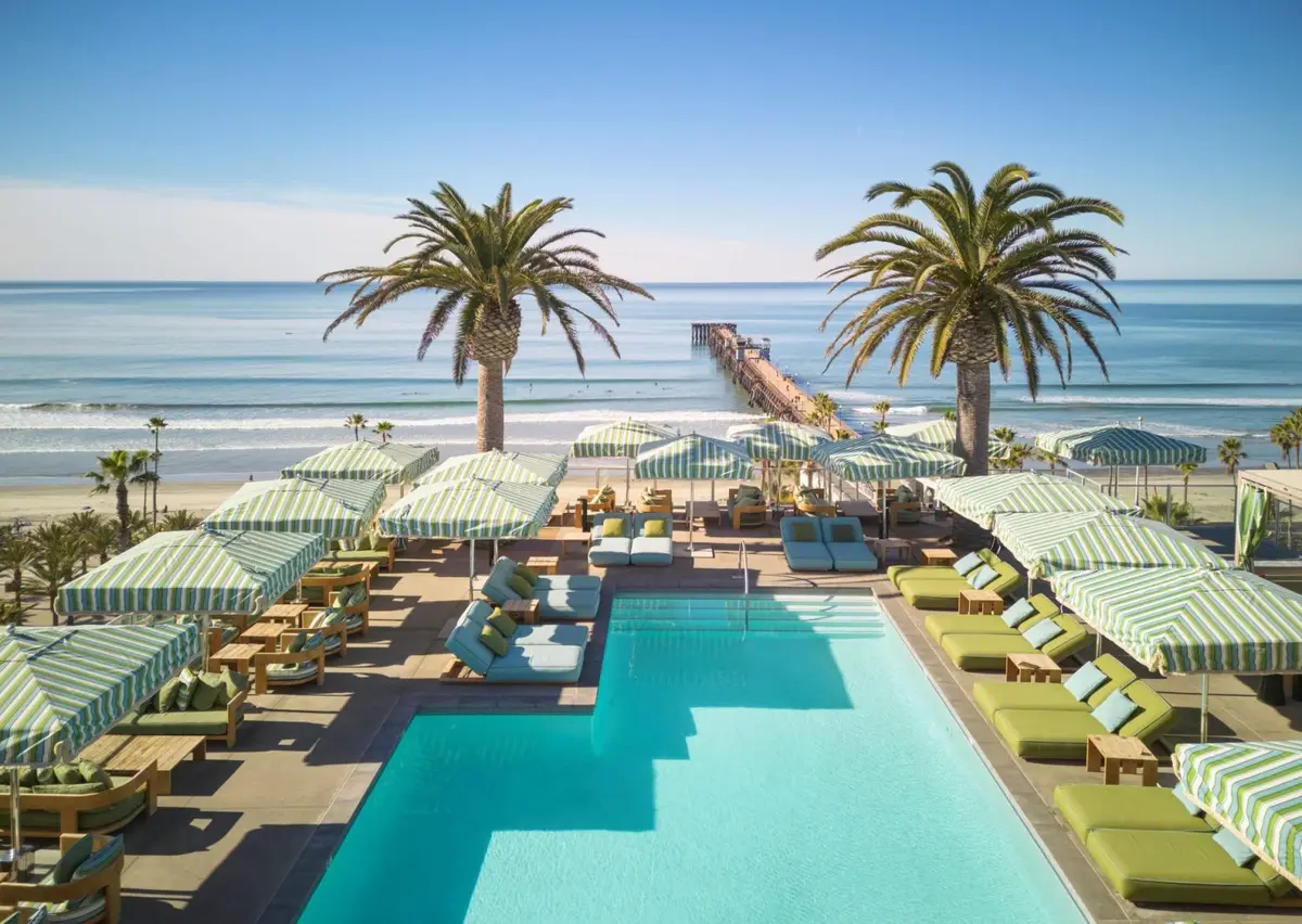 A rooftop pool from above with green striped daybeds and umbrellas, the pier and ocean behind