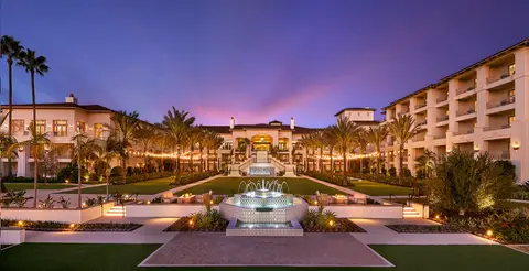 A lit Spanish colonial resort building at dusk behind a formal lawn and a tiered fountain