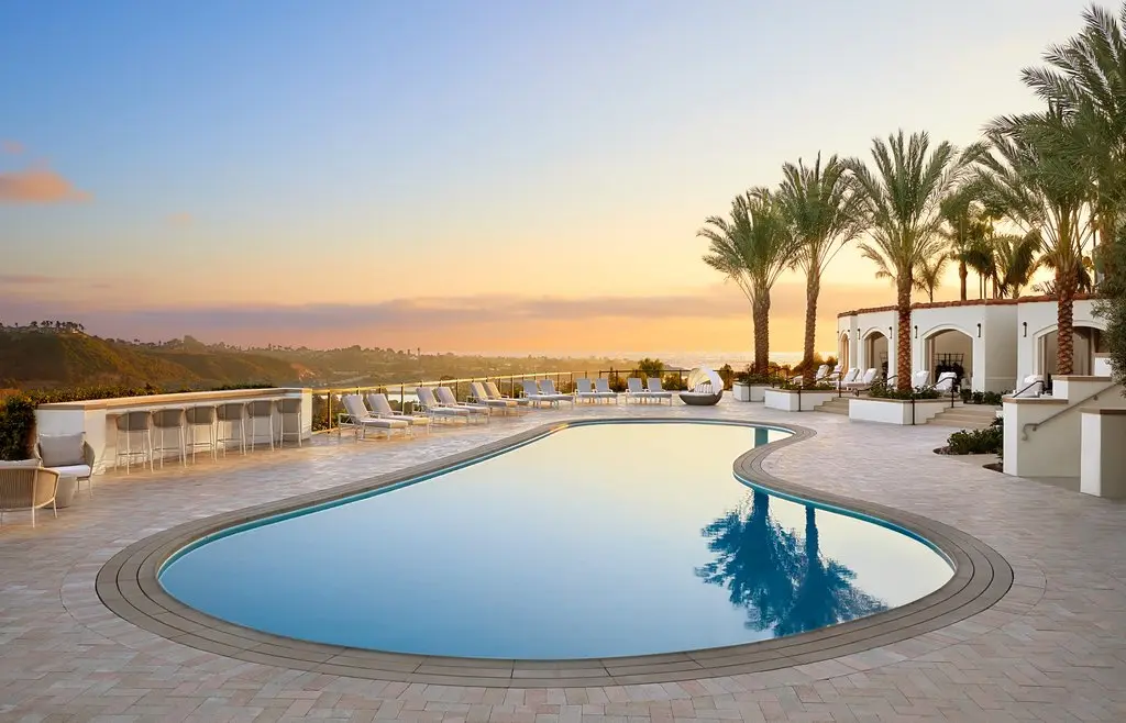 A curved pool edged with loungers and palms at sunset with a low building behind