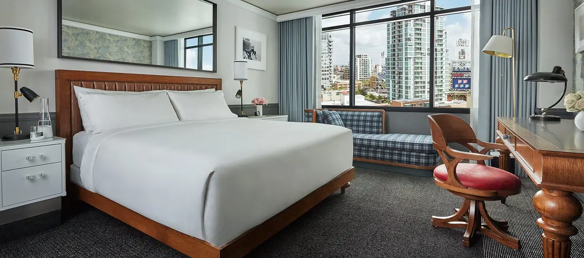 A guest room with a low bed, blue plaid window seat and floor to ceiling glass onto the city