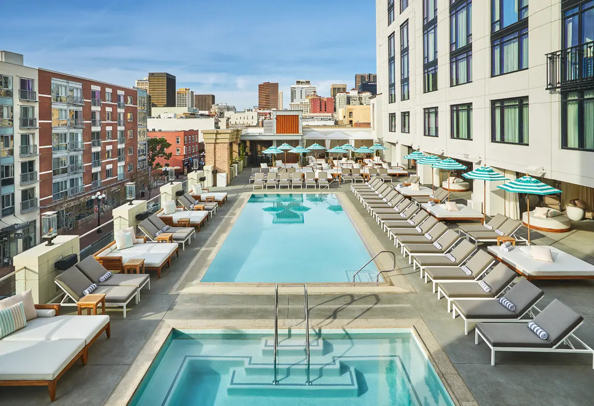 A rooftop pool flanked by loungers and turquoise umbrellas with downtown buildings behind