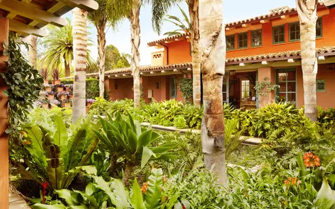 A long terracotta hacienda building with a tiled roof behind dense subtropical planting and palms