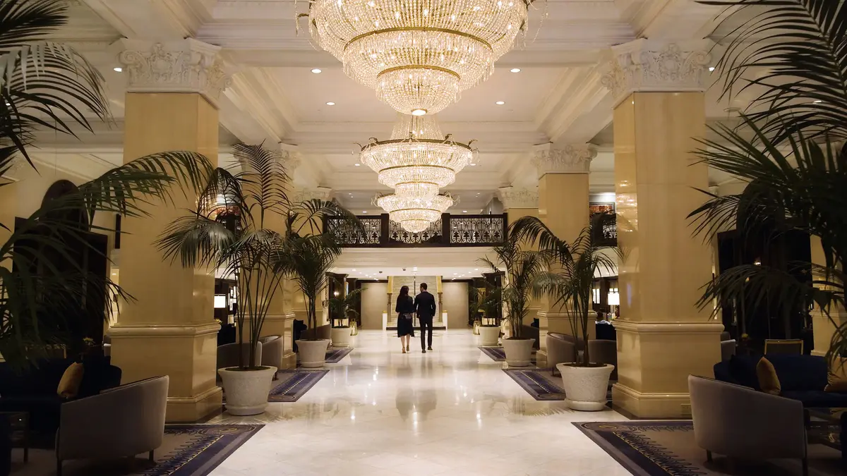 A long lobby with stone columns, chandeliers and potted palms leading to a bright doorway