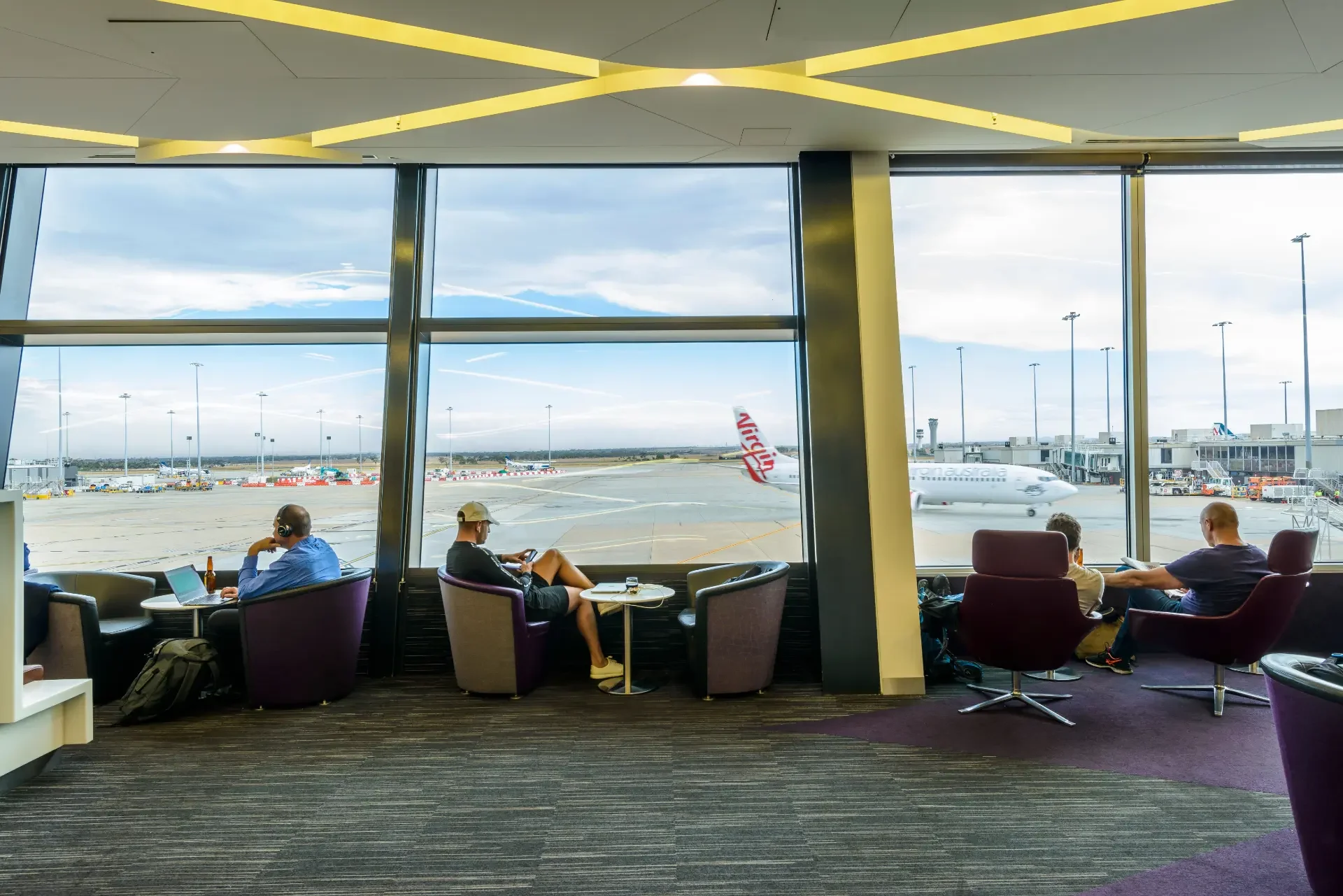 Travellers seated in an airport lounge with floor to ceiling windows overlooking the apron and an aircraft at the gate